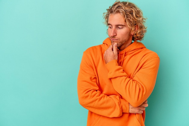 Young caucasian man isolated on blue background looking sideways with doubtful and skeptical expression.