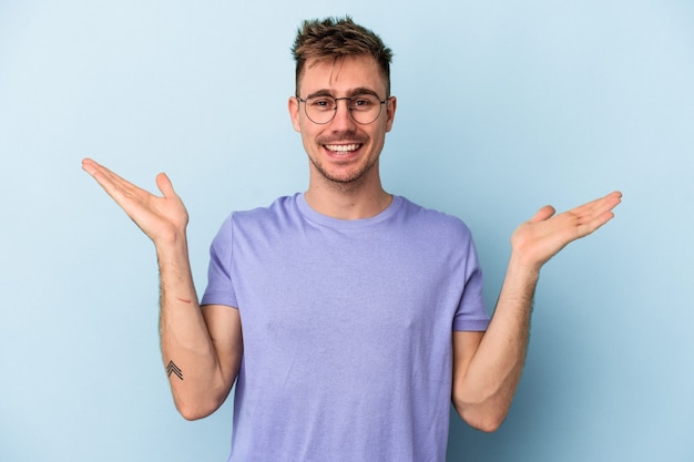 Young caucasian man isolated on blue background makes scale with arms, feels happy and confident.