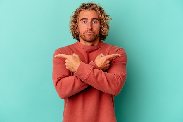 Young caucasian man isolated on blue background points sideways, is trying to choose between two options.