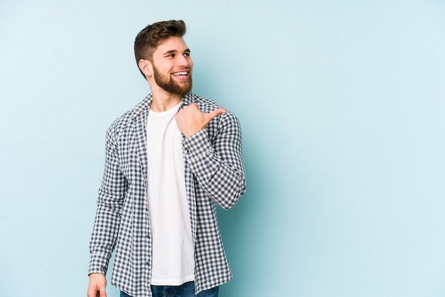 Young caucasian man isolated on blue background points with thumb finger away, laughing and carefree.