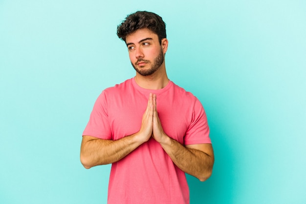 Young caucasian man isolated on blue background praying, showing devotion, religious person looking for divine inspiration.