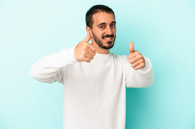 Young caucasian man isolated on blue background raising both thumbs up, smiling and confident.