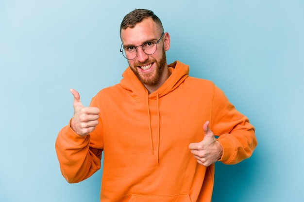 Young caucasian man isolated on blue background raising both thumbs up, smiling and confident.