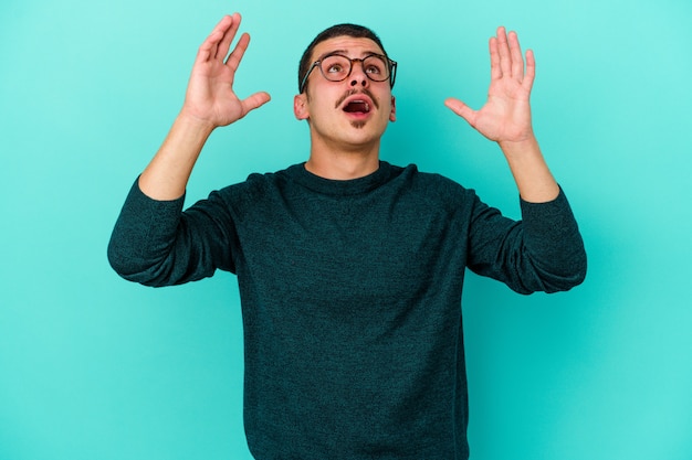 Giovane uomo caucasico isolato su sfondo blu che grida al cielo, alzando lo sguardo, frustrato.