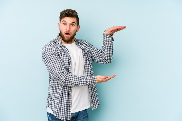 Young caucasian man isolated on blue background shocked and amazed holding a copy space between hands.