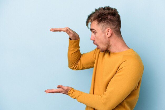 Young caucasian man isolated on blue background shocked and amazed holding a copy space between hands.