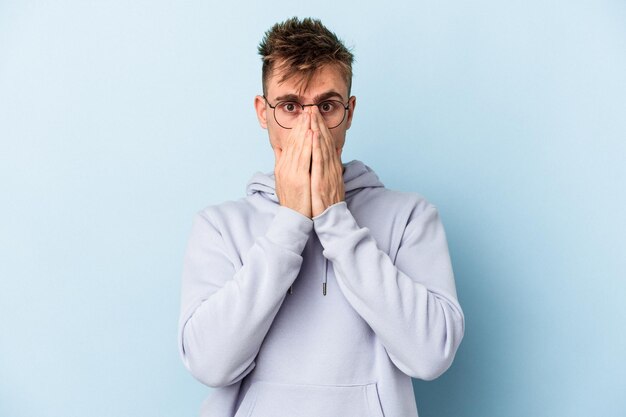Young caucasian man isolated on blue background shocked covering mouth with hands.