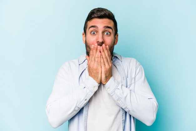 Young caucasian man isolated on blue background shocked covering mouth with hands