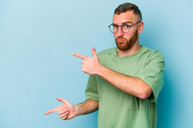 Young caucasian man isolated on blue background shocked pointing with index fingers to a copy space