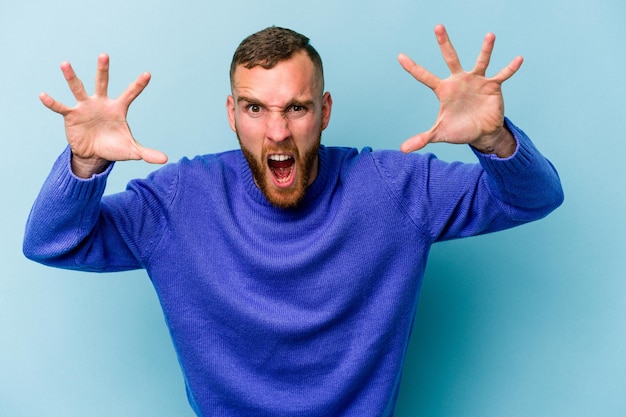 Young caucasian man isolated on blue background showing claws imitating a cat, aggressive gesture.