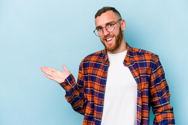 Young caucasian man isolated on blue background showing a copy space on a palm and holding another hand on waist.