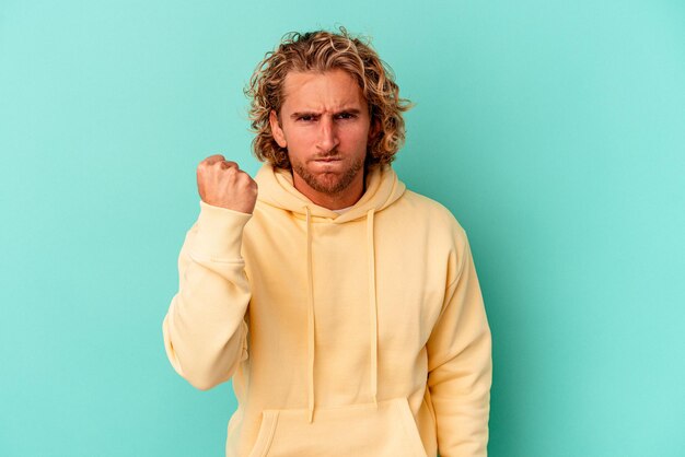 Young caucasian man isolated on blue background showing fist to camera, aggressive facial expression