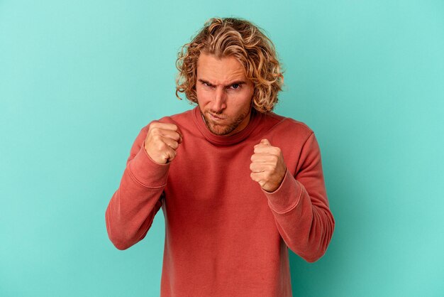 Young caucasian man isolated on blue background showing fist to camera, aggressive facial expression.