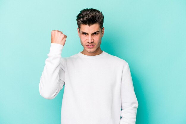 Young caucasian man isolated on blue background showing fist to camera aggressive facial expression