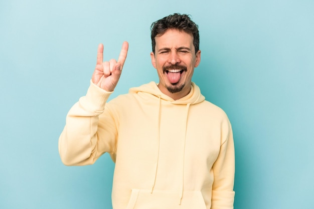 Young caucasian man isolated on blue background showing a horns gesture as a revolution concept.