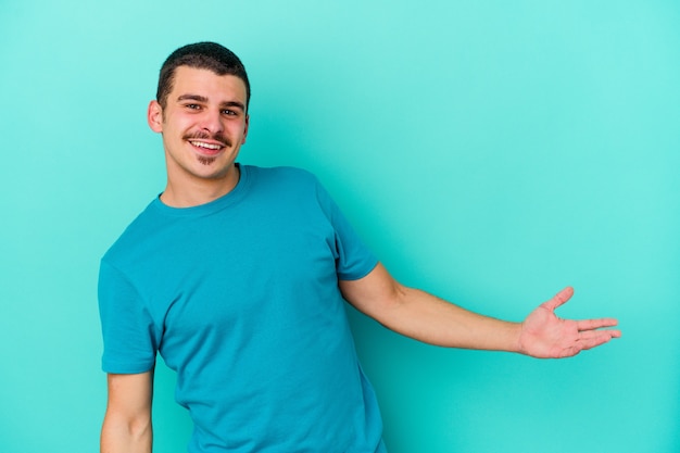 Young caucasian man isolated on blue background showing a welcome expression.