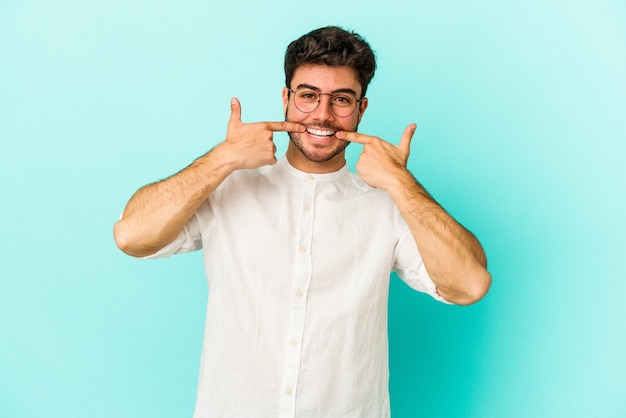 Young caucasian man isolated on blue background smiles, pointing fingers at mouth.