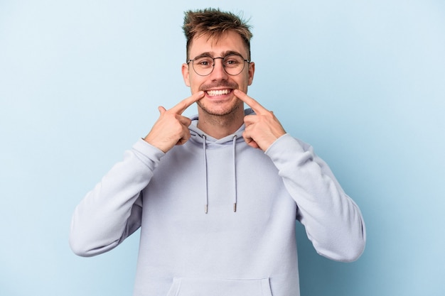 Young caucasian man isolated on blue background smiles, pointing fingers at mouth.