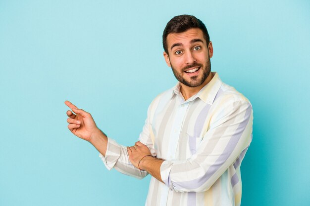 Young caucasian man isolated on blue background smiling cheerfully pointing with forefinger away.