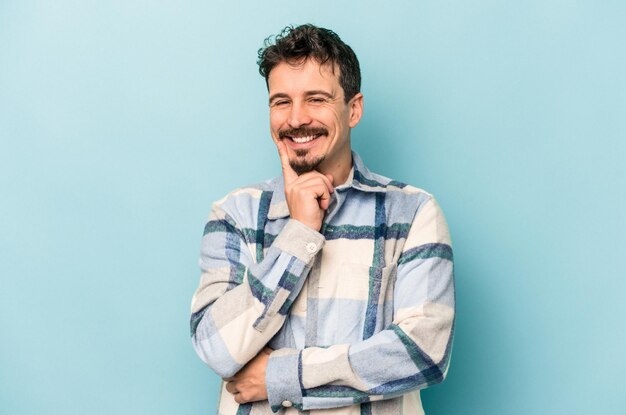 Young caucasian man isolated on blue background smiling happy and confident touching chin with hand