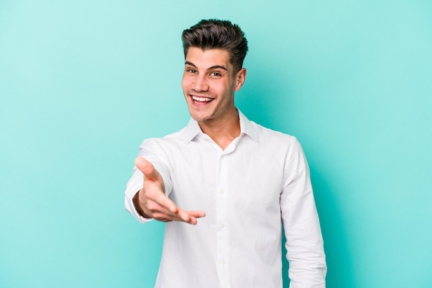 Young caucasian man isolated on blue background stretching hand at camera in greeting gesture.