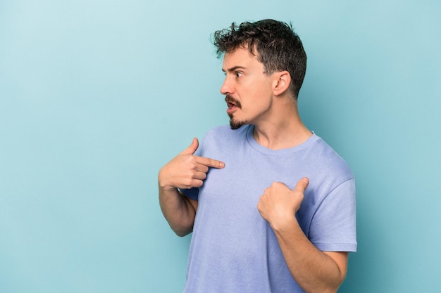 Young caucasian man isolated on blue background surprised pointing with finger, smiling broadly.