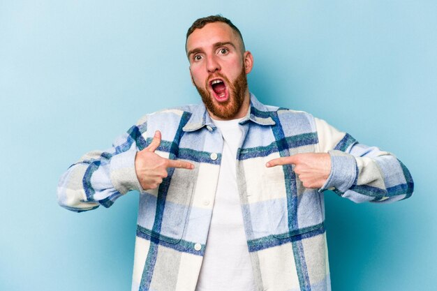 Young caucasian man isolated on blue background surprised pointing with finger, smiling broadly.