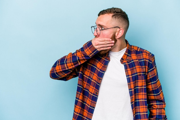 Young caucasian man isolated on blue background thoughtful looking to a copy space covering mouth with hand.