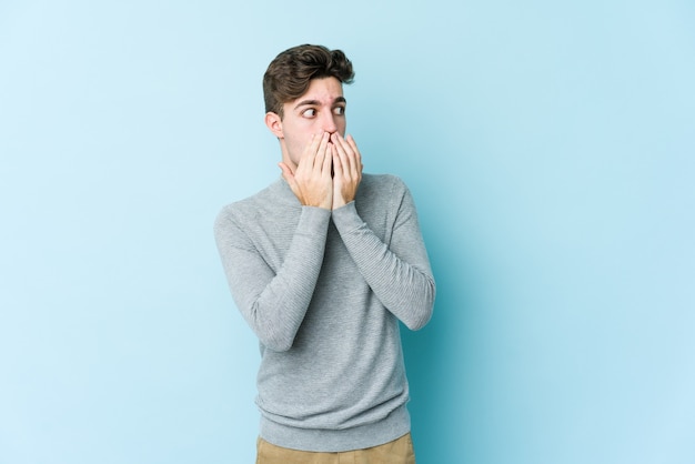 Young caucasian man isolated on blue background thoughtful looking to a copy space covering mouth with hand.