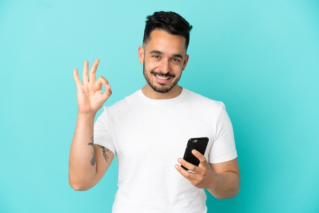 Young caucasian man isolated on blue background using mobile phone and doing OK sign