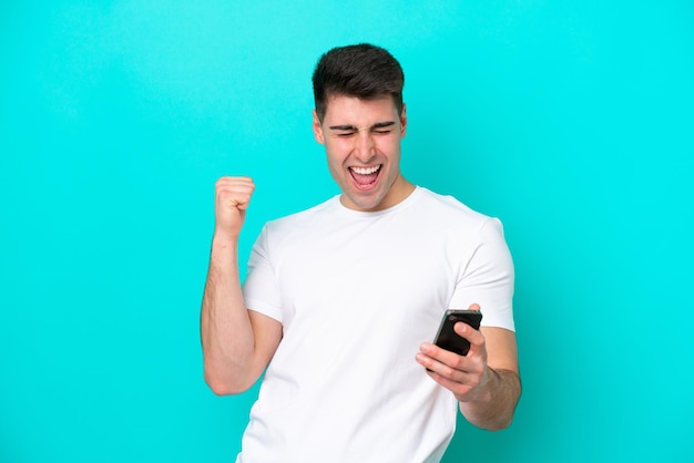 Giovane uomo caucasico isolato su sfondo blu utilizzando il telefono cellulare e facendo gesto di vittoria