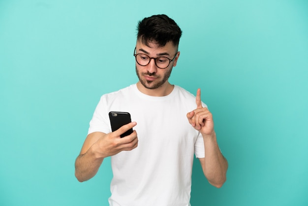 Giovane uomo caucasico isolato su sfondo blu utilizzando il telefono cellulare e sollevando il dito