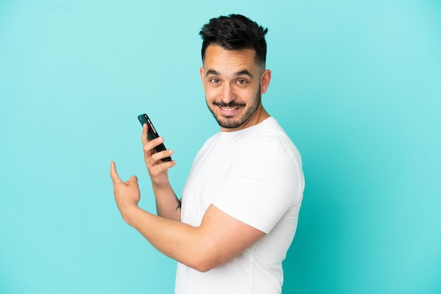 Young caucasian man isolated on blue background using mobile phone and pointing back