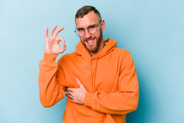 Young caucasian man isolated on blue background winks an eye and holds an okay gesture with hand