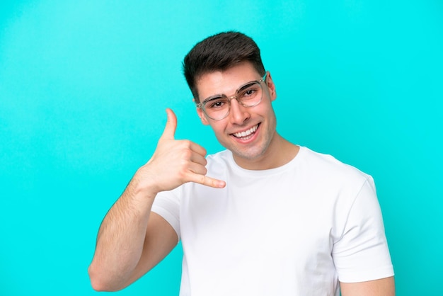 Young caucasian man isolated on blue background With glasses and doing phone gesture