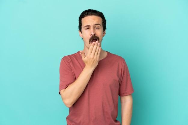 Young caucasian man isolated on blue background yawning and covering wide open mouth with hand