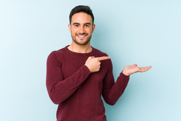 Young caucasian man isolated on blue excited holding a copy space on palm.