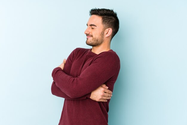 Young caucasian man isolated on blue hugs, smiling carefree and happy.