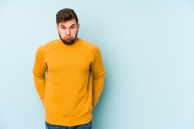 Young caucasian man isolated on blue shrugs shoulders and open eyes confused.