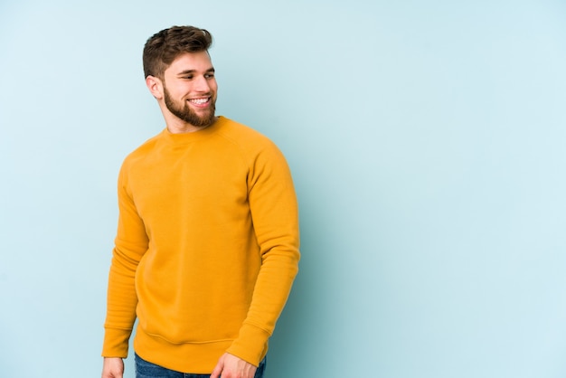 Young caucasian man isolated on blue space looks aside smiling, cheerful and pleasant.