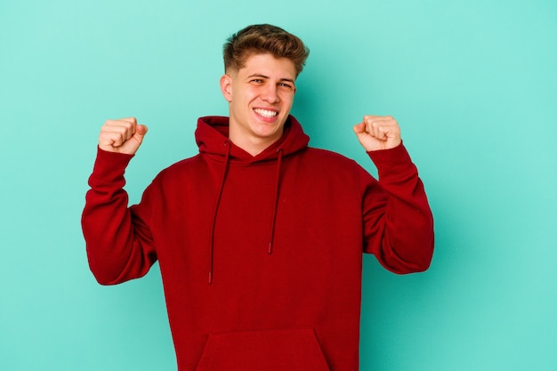 Young caucasian man isolated on blue wall cheering carefree and excited. Victory concept.