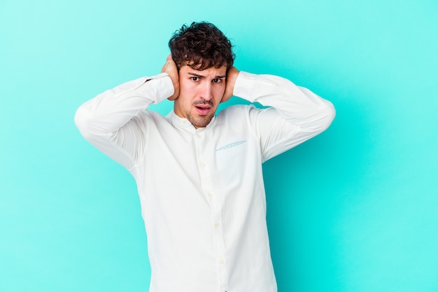 Young caucasian man isolated on blue wall covering ears with hands trying not to hear too loud sound.