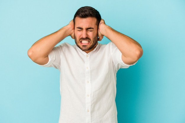 Young caucasian man isolated on blue wall covering ears with hands trying not to hear too loud sound.