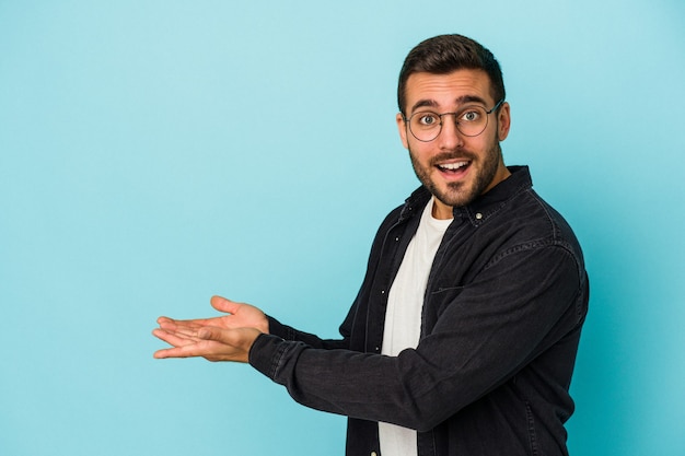 Young caucasian man isolated on blue wall holding a copy space on a palm