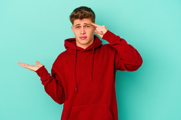 Young caucasian man isolated on blue wall holding and showing a product on hand.