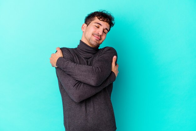 Young caucasian man isolated on blue wall hugs, smiling carefree and happy