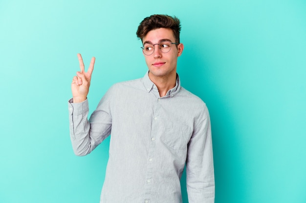 Young caucasian man isolated on blue wall joyful and carefree showing a peace symbol with fingers.