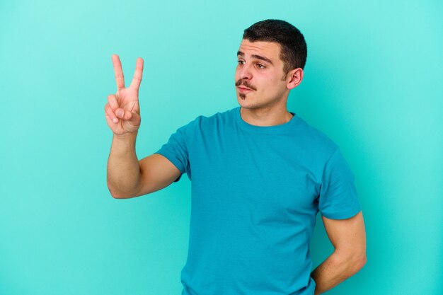 Young caucasian man isolated on blue wall joyful and carefree showing a peace symbol with fingers.