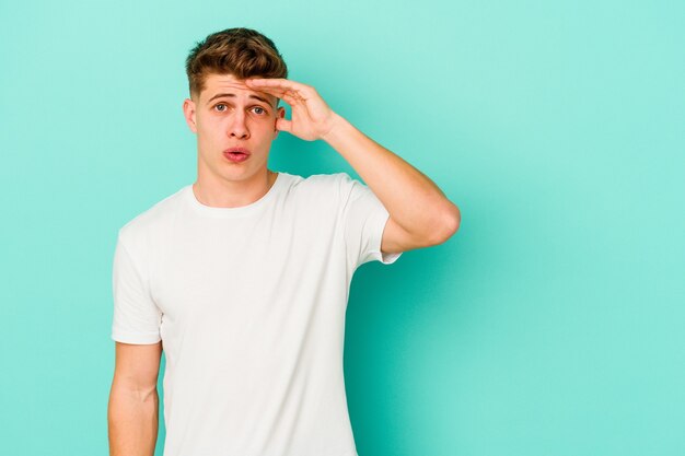 Young caucasian man isolated on blue wall looking far away keeping hand on forehead.