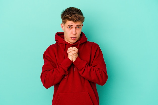 Young caucasian man isolated on blue wall praying for luck, amazed and opening mouth looking to front.